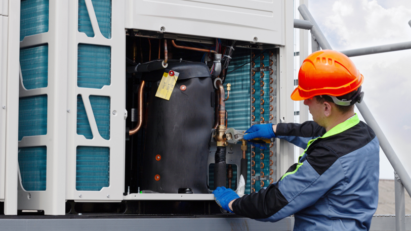 An HVAC tech in a bright orange hard hat uses a wrench on a unit.