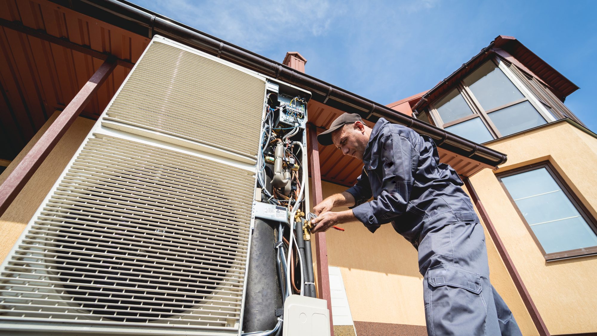 An HVAC tech uses a spanner to adjust a large unit outside of a residential building.