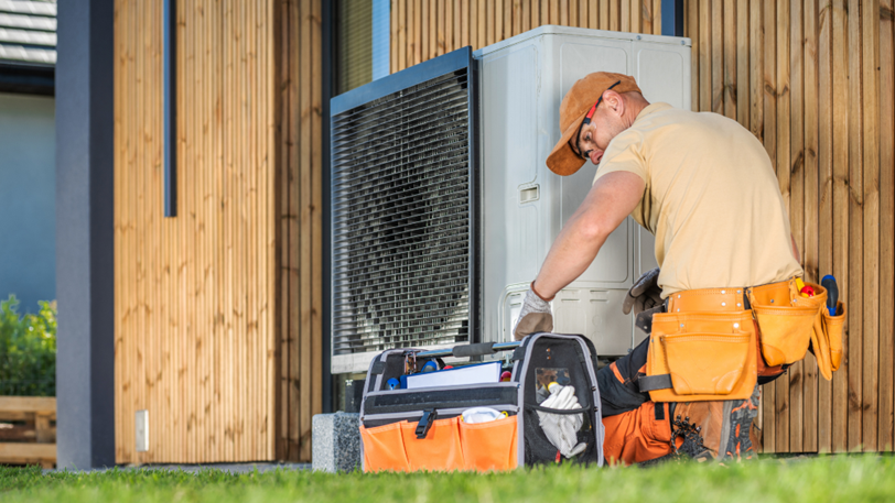 An HVAC tech wearing a ball cap, looks through a tool bag next to a unit.