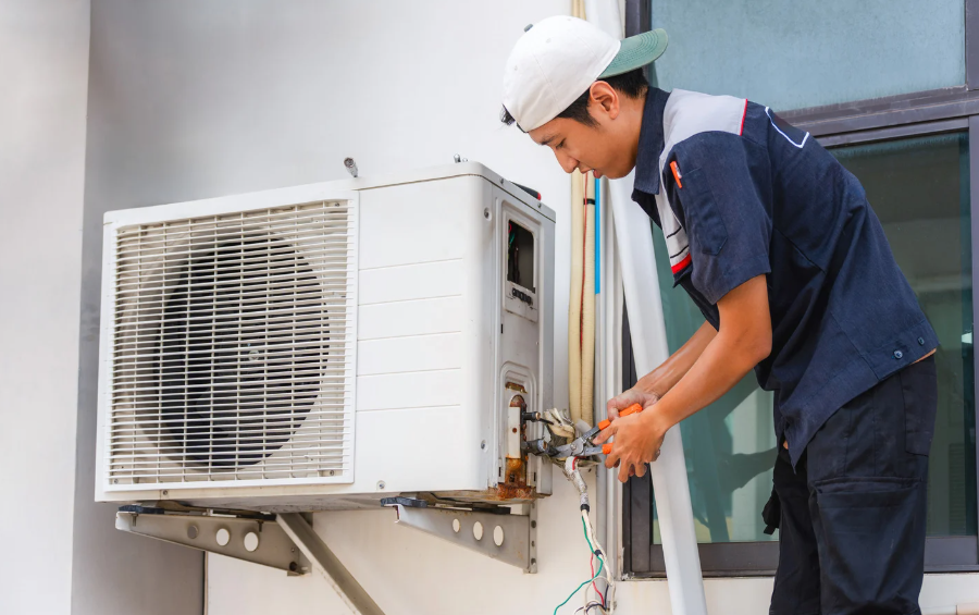 Technician in a navy uniform and white cap repairs wiring on a wall mounted white heat pump beside a dark framed window.