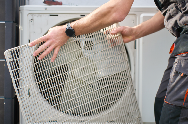Technician in dark workwear removes the white grid-patterned cover from a heat pump unit, exposing the large circular fan inside.