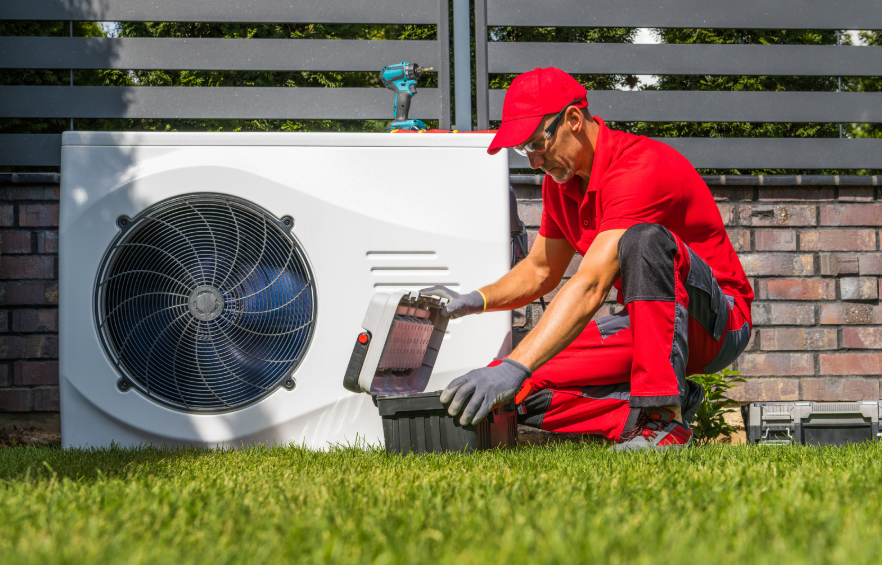 Technician in a bright red uniform services a white outdoor HVAC unit with a black circular fan beside a brick wall and green lawn.